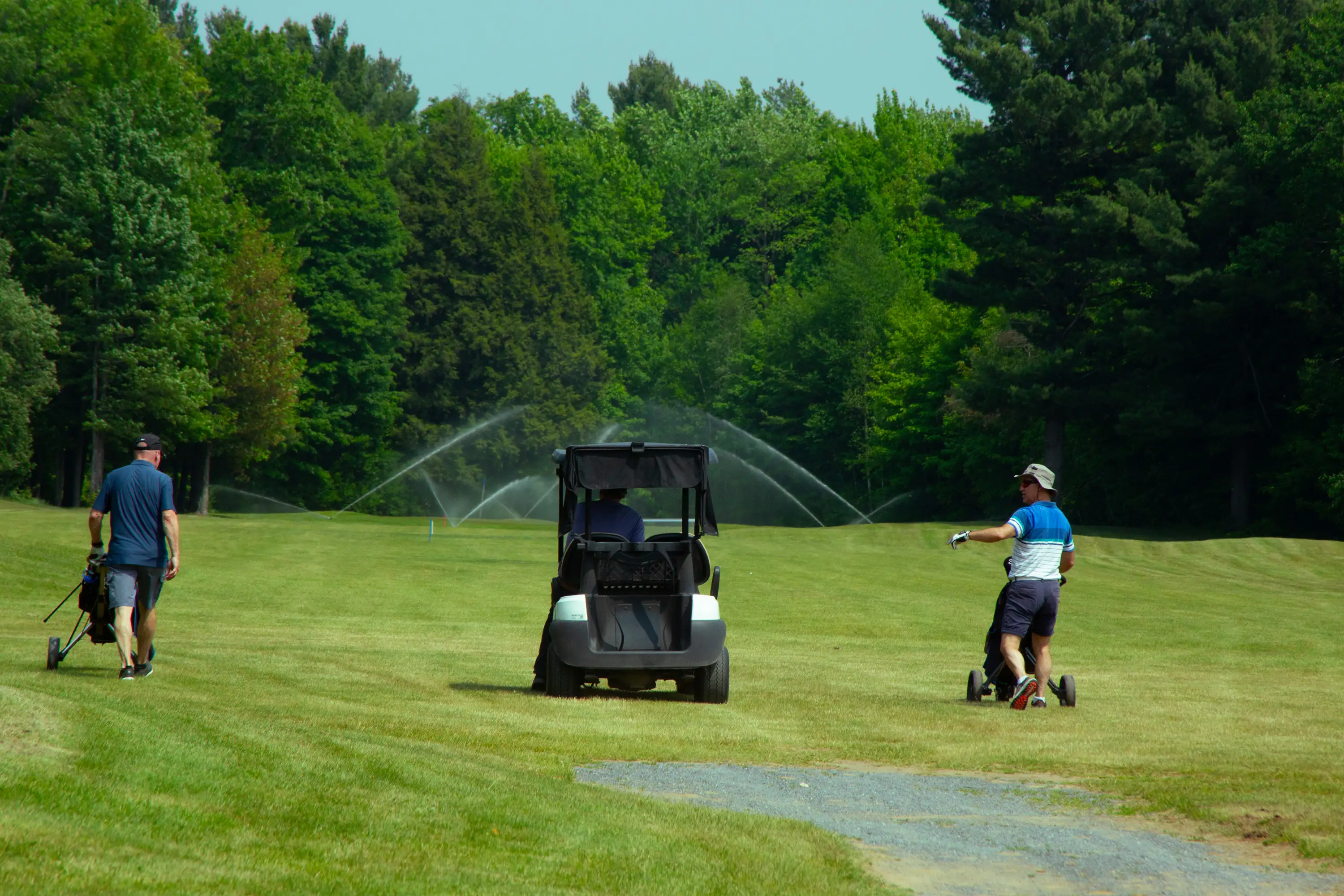 Joueurs sur un fairway ensoleillé avec voiturette et arrosage en arrière-plan — tournoi de golf aux Patriotes